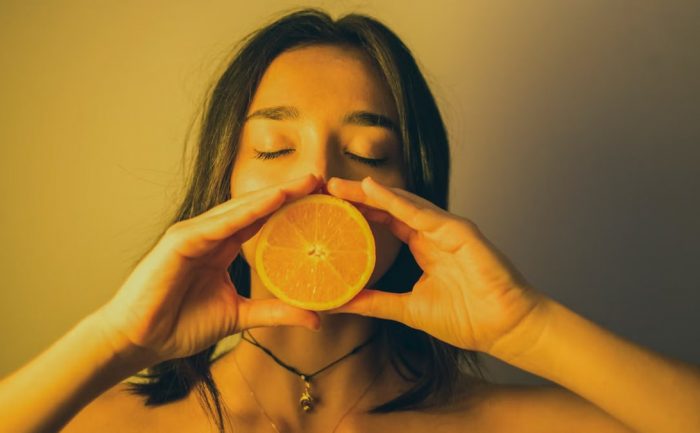 Calm young female with bare shoulders standing with eyes closed against plain wall and covering lips with orange slice