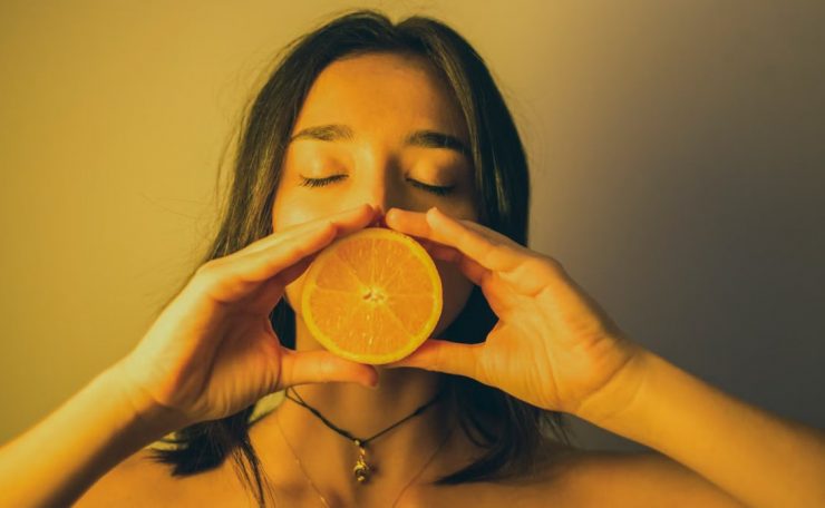 Calm young female with bare shoulders standing with eyes closed against plain wall and covering lips with orange slice